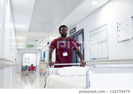 African American male hospital worker pushing bed in hospital corridor, with IV pole and ID badge 128738479
