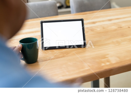 Man sitting at wooden table at home, holding green mug while viewing tablet screen Man sitting at wooden table at home, holding green mug while viewing tablet screen 128738480