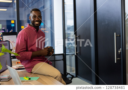 African American man sitting on desk in office, holding coffee mug with open laptop notebook 128738481
