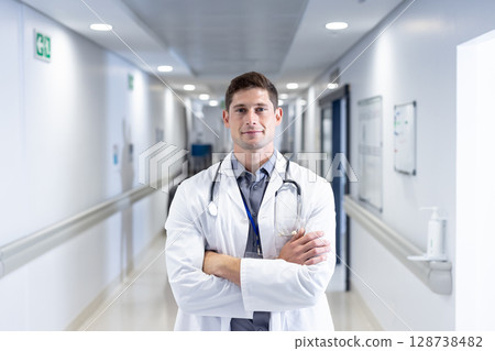 Young male doctor standing in hospital corridor, wearing lab coat with stethoscope and ID badge 128738482