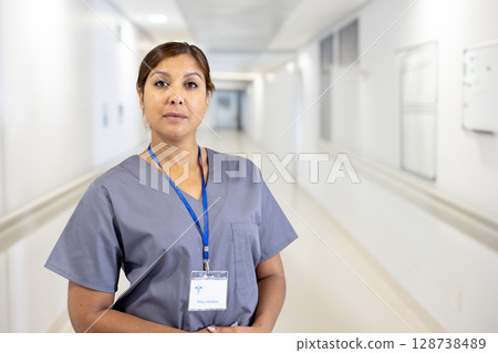 Senior woman standing in bright hospital corridor, wearing gray scrubs and blue lanyard badge 128738489