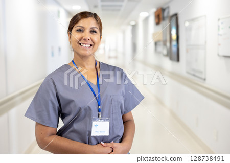 Diverse woman wearing scrubs and ID badge, standing in hospital corridor with whiteboard, smiling 128738491