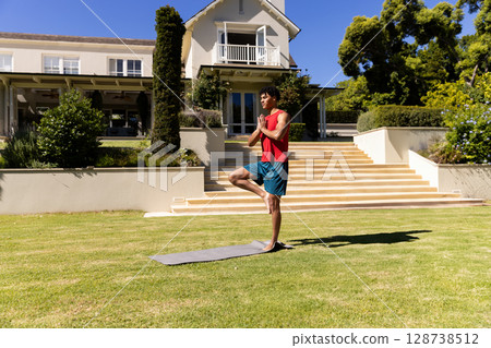 Young man practicing tree pose on lawn in front of two-story house, with gray yoga mat 128738512