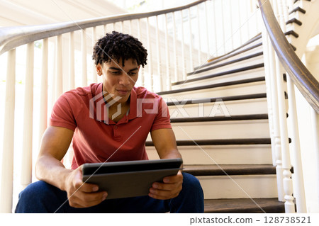 Man in early 20s sitting on curved dark wood staircase in stairwell, using tablet computer Man in early 20s sitting on curved dark wood staircase in stairwell, using tablet computer 128738521