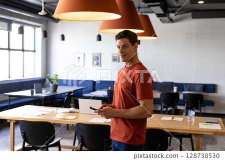 Mid adult man standing at wooden table in open-plan office, using smartphone and smiling 128738530