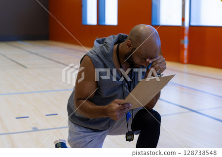 Mid-adult male coach kneeling on indoor basketball court, wiping sweat with clipboard and stopwatch Mid-adult male coach kneeling on indoor basketball court, wiping sweat with clipboard and stopwatch 128738548