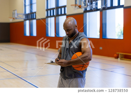 male coach standing on indoor basketball court, holding basketball and noting on clipboard with pen 128738550