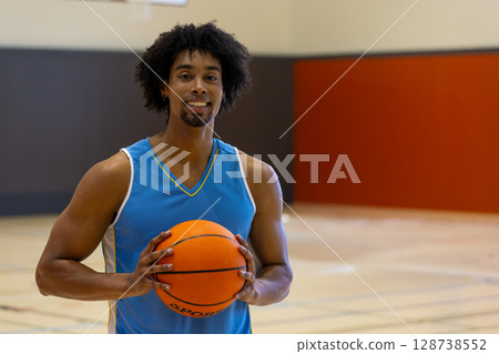 African American man standing on gym basketball court, holding basketball and smiling at camera African American man standing on gym basketball court, holding basketball and smiling at camera 128738552