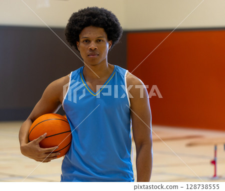 Young African American man standing on gymnasium court, holding orange basketball under arm 128738555