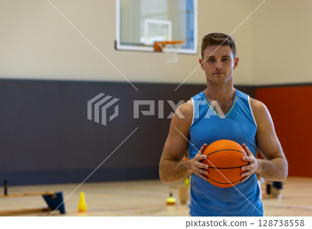 Male basketball player holding basketball on gym court, with training cones and hoop, copy space 128738558