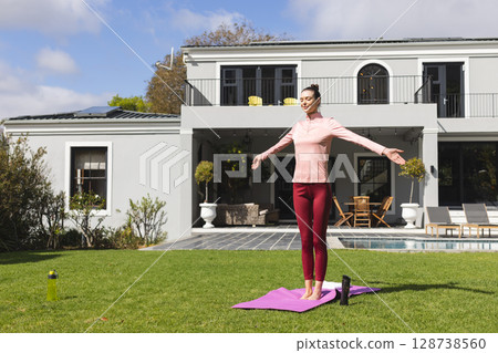 Woman performing yoga stretch on pink mat on green lawn in suburban backyard, with water bottles Woman performing yoga stretch on pink mat on green lawn in suburban backyard, with water bottles 128738560