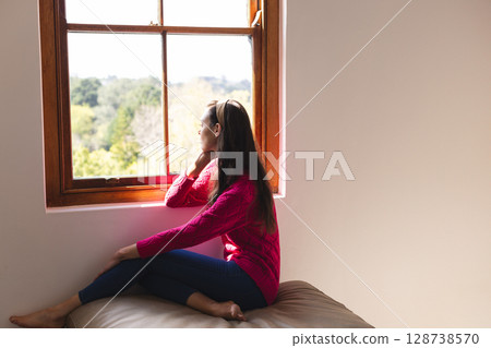 Woman sitting on window seat indoors, gazing thoughtfully out wooden-framed window, copy space 128738570