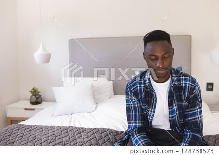 Young African American man sitting on bed edge in modern bedroom, with grey quilt, copy space 128738573