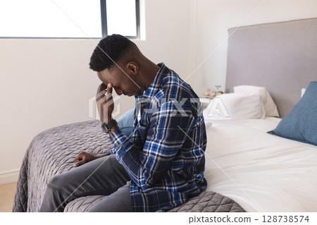 African American man sitting on bed in sunlit bedroom with window, pinching nose, copy space 128738574