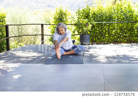 Senior woman sitting on dark exercise mat on deck stretching forward toward foot, with potted plant 128738578