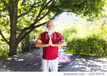 Senior man praying on wooden deck under tree canopy with rope railing and pink yoga mat 128738579