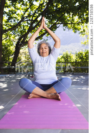 Senior woman practicing yoga by metal railing on terrace, using pink yoga mat with water bottle 128738586