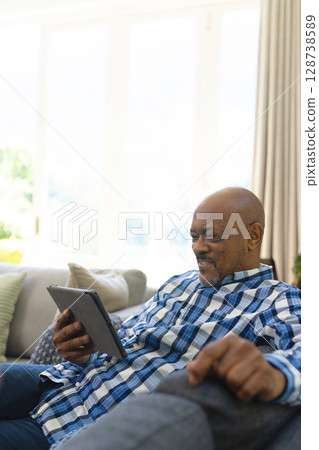 Senior man sitting on grey sofa in bright living room, holding tablet with cushions, copy space 128738589