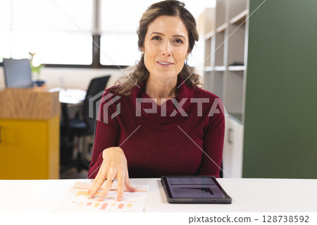 Woman analyzing printed bar graphs on white desk in modern office, using tablet and reviewing data Woman analyzing printed bar graphs on white desk in modern office, using tablet and reviewing data 128738592