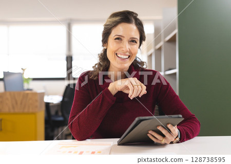 Businesswoman sitting at desk in bright modern office, holding tablet and displaying bar charts 128738595