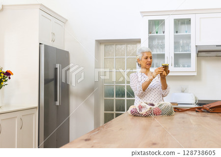Senior woman holding jar behind island in modern home kitchen, with mesh produce bag, copy space Senior woman holding jar behind island in modern home kitchen, with mesh produce bag, copy space 128738605