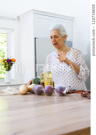 Senior woman standing behind island in kitchen, examining fresh produce with knife, copy space 128738608