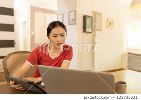 Young Asian woman leaning at wooden table in home, using laptop and tablet with coffee mug 128738621