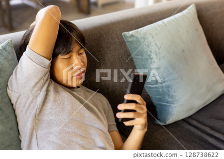 Young Asian man reclining on gray couch at home with teal pillows, using smartphone, copy space 128738622