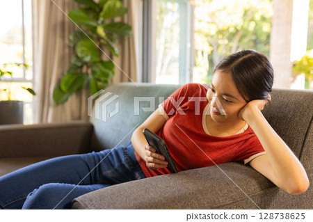 Asian woman reclining on grey sofa in living room with potted plant, holding smartphone, copy space 128738625