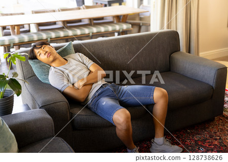 Asian man reclining on dark gray sofa in living room, with teal pillow, rug, striped bench 128738626