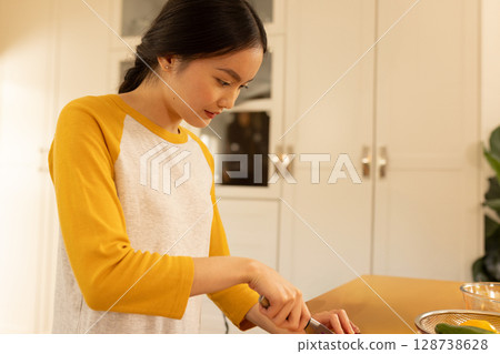Asian woman slicing cucumber on wooden cutting board at home kitchen island, with glass bowls Asian woman slicing cucumber on wooden cutting board at home kitchen island, with glass bowls 128738628