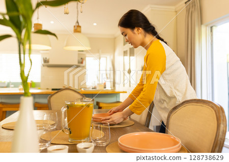 Asian woman arranging tableware on dining table in kitchen area, featuring citrus juice pitcher Asian woman arranging tableware on dining table in kitchen area, featuring citrus juice pitcher 128738629