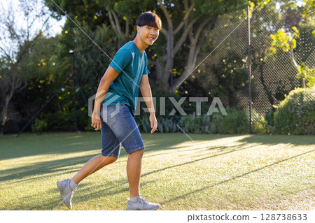 Young Asian man walking across synthetic turf in backyard, with chain-link fence, shrubs and trees 128738633