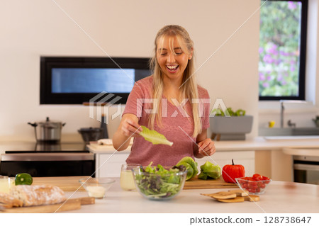 Young woman preparing salad in glass bowl on island in modern kitchen, with bread and tomatoes 128738647