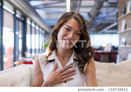Young adult woman placing hand on chest and smiling sitting on beige couch in office lounge 128738652