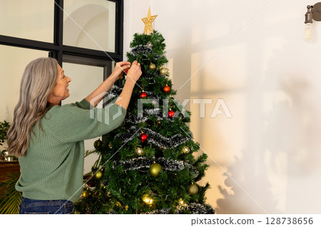 Senior woman placing red ornament on Christmas tree in modern living room with gold baubles 128738656