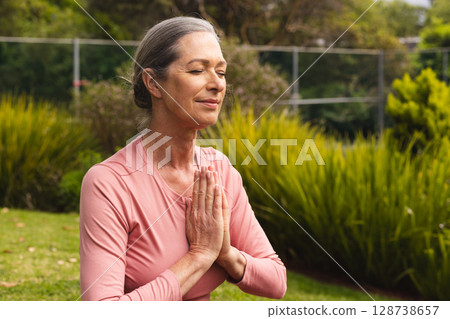 Senior female kneeling on grassy lawn in backyard garden, meditating with eyes closed 128738657