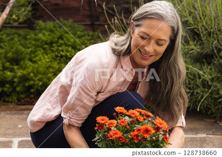 Senior woman kneeling while tending potted orange mums on backyard patio, enjoying gardening 128738660