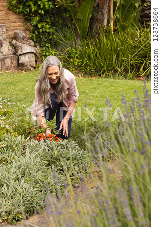 Senior woman kneeling and tending orange flowers in backyard garden, with lavender and shrubs 128738664