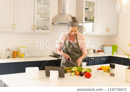 Chopping senior woman preparing vegetables on wooden cutting board on kitchen island, with tablet 128738672