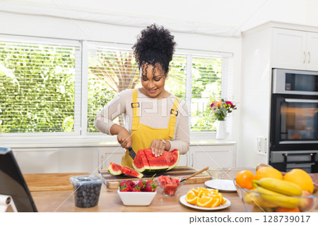 Woman slicing watermelon on cutting board on island in kitchen, with fruit bowl and tablet Woman slicing watermelon on cutting board on island in kitchen, with fruit bowl and tablet 128739017