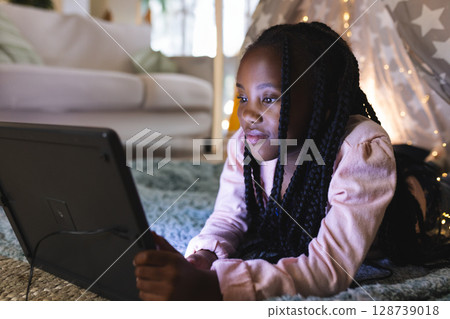 African American girl lying on stomach using tablet at home, star tent, string lights, copy space African American girl lying on stomach using tablet at home, star tent, string lights, copy space 128739018