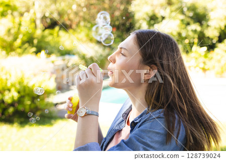 Woman in twenties blowing soap bubbles with bubble wand in backyard near pool, copy space 128739024