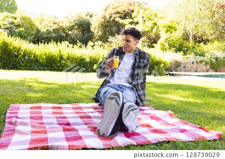 Man sitting on red white checkered picnic blanket in backyard, holding glass of orange juice Man sitting on red white checkered picnic blanket in backyard, holding glass of orange juice 128739029