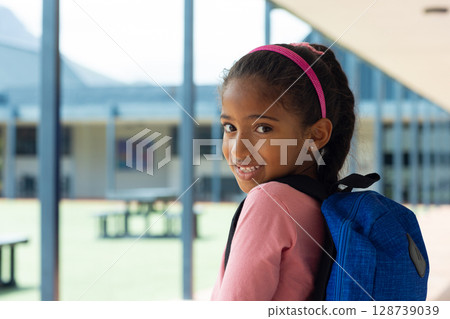 Multiracial girl child standing under covered school walkway, holding blue backpack, copy space 128739039