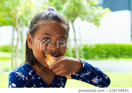 Young girl sitting outdoors in green backyard, holding torn bread piece and smiling Young girl sitting outdoors in green backyard, holding torn bread piece and smiling 128739041
