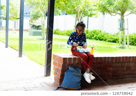 Child girl eating lunch on brick wall in courtyard, with tray, bottle and backpack, copy space Child girl eating lunch on brick wall in courtyard, with tray, bottle and backpack, copy space 128739042