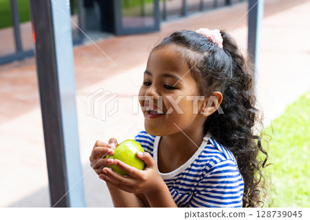 Multiracial female child holding green apple while sitting on tiled patio near glass door entrance Multiracial female child holding green apple while sitting on tiled patio near glass door entrance 128739045