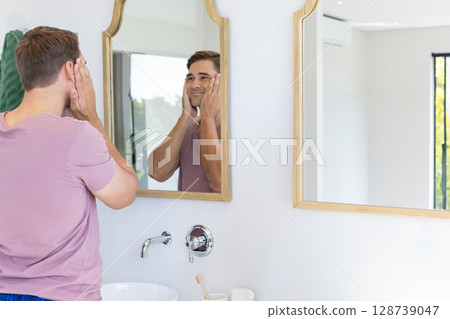 Man touching face at sink mirror in bright modern bathroom, with toothbrush and towel, copy space 128739047