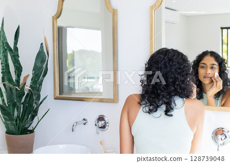 Asian Indian woman applying facial pad in home bathroom with wooden mirror and plant, copy space Asian Indian woman applying facial pad in home bathroom with wooden mirror and plant, copy space 128739048
